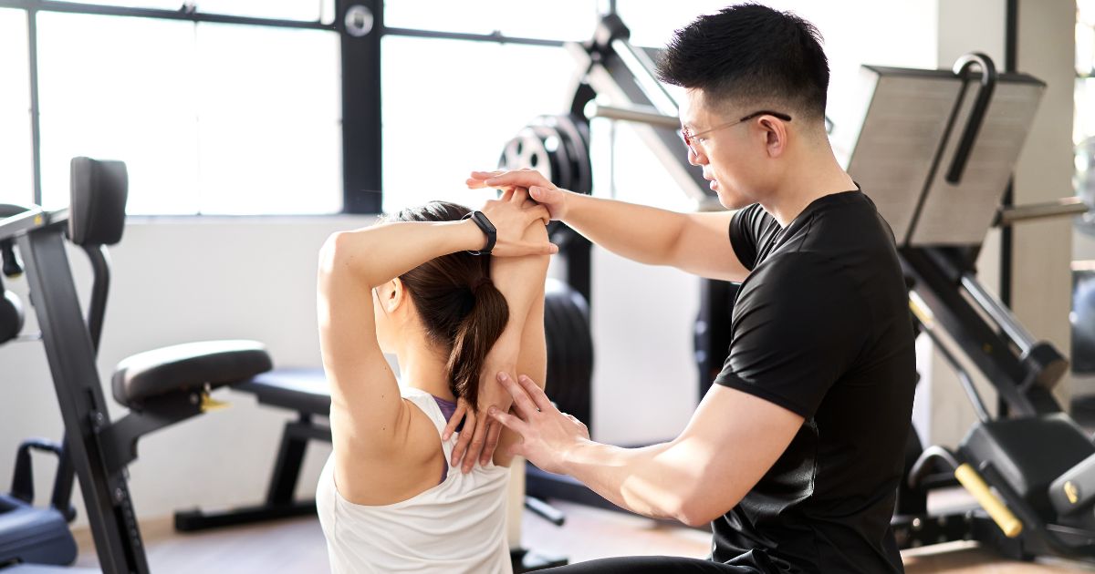 ストレッチを受けている女性が、トレーナーによって腕後ろ周りを伸ばされている様子。